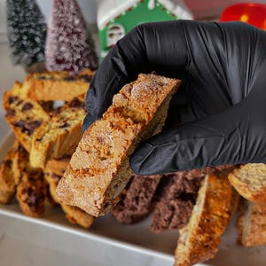 May include: A close-up of a hand holding a light brown biscotti cookie. The cookie is slightly dusted with powdered sugar. The cookie is surrounded by other biscotti cookies, some of which are chocolate flavored.