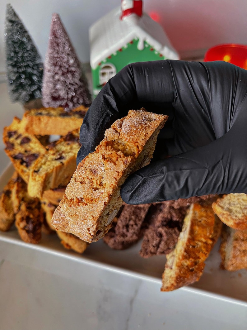 May include: A close-up of a hand holding a light brown biscotti cookie. The cookie is slightly dusted with powdered sugar. The cookie is surrounded by other biscotti cookies, some of which are chocolate flavored.