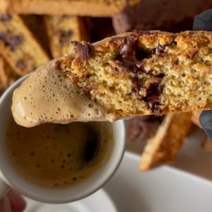 May include: A close-up of a biscotti cookie dipped in a cup of coffee. The biscotti is light brown with chocolate chips and the coffee is light brown with foam.