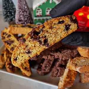 May include: A close-up of a broken biscotti cookie with chocolate chips. The cookie is light brown and has a crispy texture. The cookie is held in a gloved hand. The cookie is surrounded by other biscotti cookies, some of which are chocolate flavored.