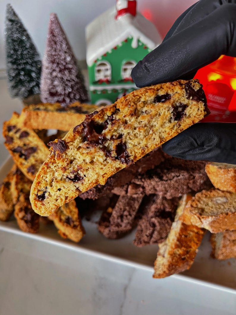 May include: A close-up of a broken biscotti cookie with chocolate chips. The cookie is light brown and has a crispy texture. The cookie is held in a gloved hand. The cookie is surrounded by other biscotti cookies, some of which are chocolate flavored.