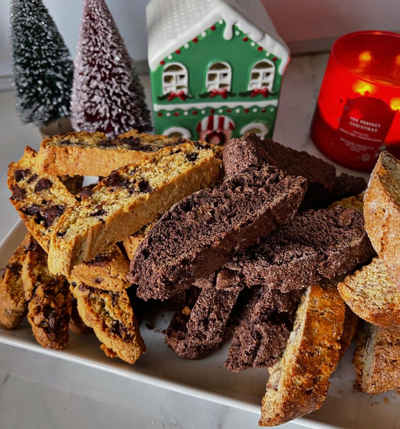 May include: A white platter with a pile of biscotti. The biscotti are a mix of chocolate and plain. Some of the biscotti have chocolate chips. The biscotti are arranged in a pile on the platter. There is a small green house and a red candle in the background.