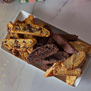 May include: A white rectangular plate with a variety of biscotti. The biscotti are a mix of chocolate chip and almond biscotti. The biscotti are arranged in a pile on the plate.