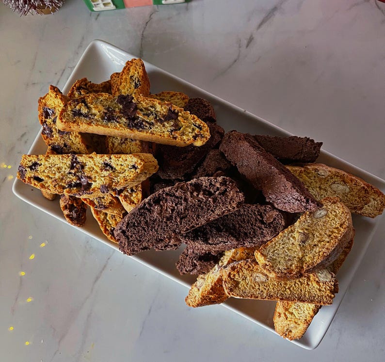 May include: A white rectangular plate with a variety of biscotti. The biscotti are a mix of chocolate chip and almond biscotti. The biscotti are arranged in a pile on the plate.