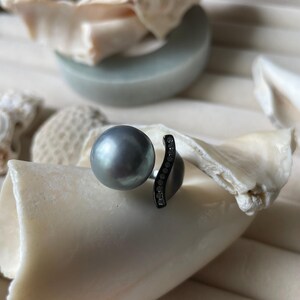 May include: A gray pearl ring with a dark metal band. The ring features a large, round pearl and a curved band adorned with small, clear stones. The ring is displayed on a white, textured surface, suggesting a jewelry or accessory theme.