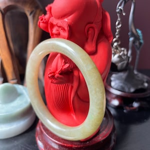 May include: A jade green bangle bracelet with a light green hue, resting on a red figurine of a smiling man. The figurine is on a wooden stand.