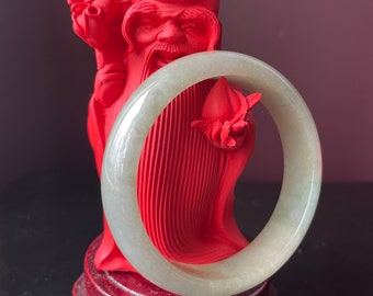 May include: A red figurine of a Chinese deity holding a jade green bangle. The deity is wearing a red robe and has a long white beard. The figurine is standing on a wooden base.