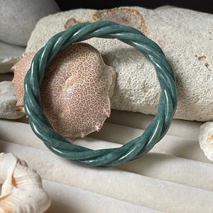 May include: A circular bangle bracelet crafted from smooth, polished green stone. The bangle features a continuous twisted rope design, showcasing deep forest green hues with subtle variations in color and a glossy, reflective surface finish.