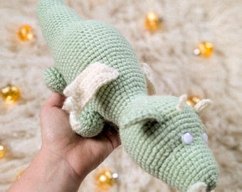 May include: A light green crocheted dragon toy with white accents on the wings and beard. The dragon has white eyes and small horns. The toy is made of soft yarn and is a handmade item.