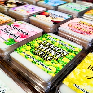 May include: A collection of square soy wax melts in clear plastic containers, each featuring a colorful label with text such as Lemon Mint Leaf, Hawaiian Hibiscus, Life's A Peach, and Honey Jasmine, showcasing various fruit and floral illustrations.