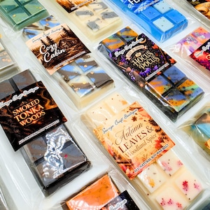 May include: A collection of rectangular soy wax melt bars in clear plastic packaging, each featuring a unique label with names like Smoked Tonka Woods, Autumn Leaves & Woodland Spice, Cozy Cabin, Hocus Pocus, and Fall Magic, with varied colors.