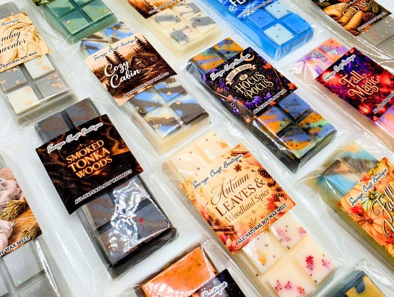 May include: A collection of rectangular soy wax melt bars in clear plastic packaging, each featuring a unique label with names like Smoked Tonka Woods, Autumn Leaves & Woodland Spice, Cozy Cabin, Hocus Pocus, and Fall Magic, with varied colors.