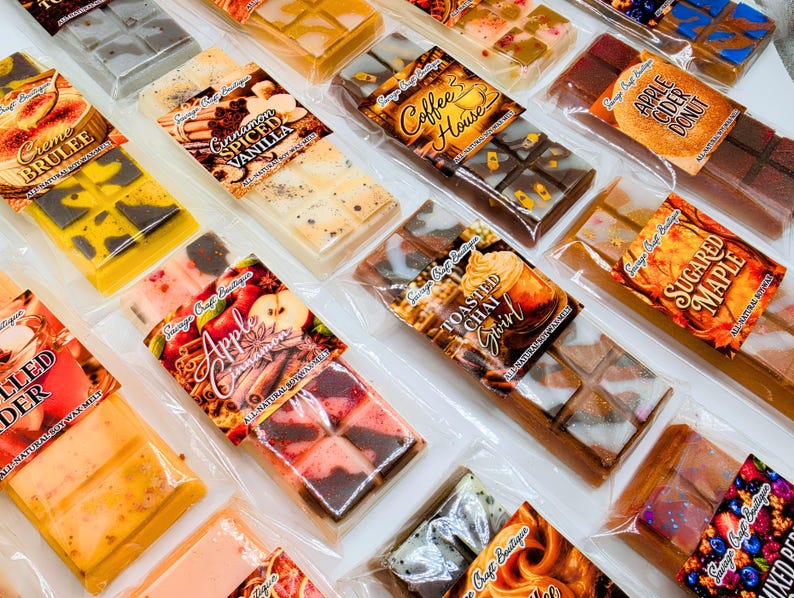 May include: A collection of rectangular soy wax melt bars in clear plastic packaging, each featuring a unique label with scent names like Creme Brulee, Cinnamon Spiced Vanilla, Coffee House, Apple Cider Donut, Toasted Chai, and Sugared Maple.