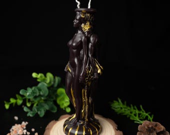 May include: A dark brown candle shaped like a three-faced female figure, with gold accents. The candle sits on a wooden slice, with a white wick extending from the top. The background is black, with greenery and spices.