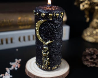 May include: A black pillar candle with a gold crescent moon and skeleton design. The candle has a textured surface and is adorned with gold skulls. It sits on a wooden slice and is lit with a flame.