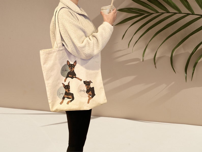 Pu&ograve; includere: Una borsa tote in tela bianca con tre illustrazioni di cani neri e marroni. I cani si chiamano "Baby", "Louis" e "Shady".
