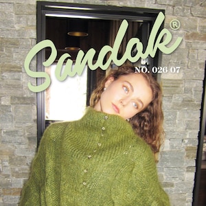 May include: A woman wearing a green knit sweater with a high collar and button details. The sweater is part of a magazine cover with the word "Sandak" in a light green script. The text "NO. 026 07" is also visible.