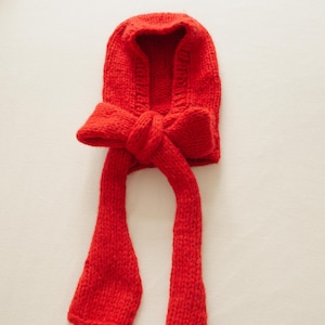 Könnte beinhalten: Eine leuchtend rote Strickhaube mit einer großen Schleife vorne und langen, herabhängenden Bändern. Die Haube ist aus einem dicken, strukturierten Garn gefertigt, wobei die Schleife ein auffälliges Merkmal ist. Die Bänder hängen herab und verleihen dem Design einen verspielten Touch.