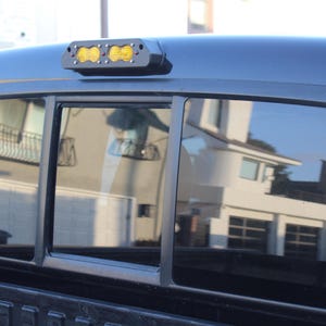 May include: Black truck cab with a rear window and a black light bar with two yellow lights. The window reflects a house and the sky. The truck bed is visible.