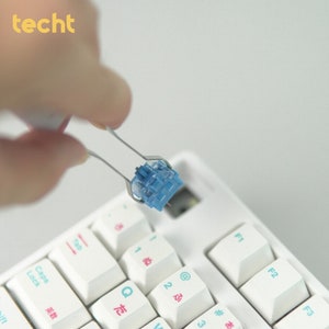 May include: A close-up of a blue mechanical keyboard switch being held by tweezers above a white keyboard. The keyboard has white keys with black lettering and pink and blue accents.