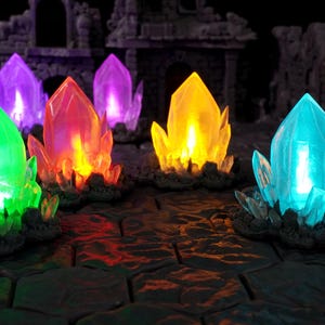 Pode incluir: Uma coleção de seis formações de cristal translúcidas e multifacetadas em tons vibrantes de verde, roxo, laranja, amarelo e azul. Cada cristal é montado sobre uma base rochosa escura e texturizada e emite um brilho interno intenso.