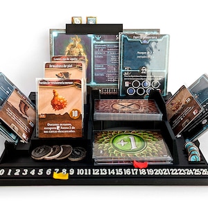 Puede incluir: Un organizador de juegos de mesa negro con múltiples compartimentos para cartas, fichas y piezas de juego. El organizador tiene una base numerada del 0 al 26. Se ven varias cartas de juego con ilustraciones y texto, junto con fichas y dados.