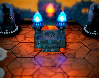 Pode incluir: Um altar de pedra em miniatura com duas tochas de chama azul no topo. O altar está em um tabuleiro de jogo hexagonal com um padrão vermelho e laranja.