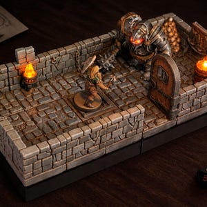 Op de afbeelding: Een miniatuur kerker-diorama met grijze stenen muren en vloertegels, een houten deur en twee oplichtende oranje LED-fakkels. Een krijgersfiguur staat tegenover een groot gepantserd monster met gloeiende rode ogen. Drie dobbelstenen liggen ernaast.