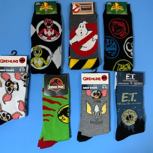 以下が含まれることがあります： ポップカルチャーのキャラクターやロゴが描かれたカラフルなクルーソックス10足セット。青い背景にディスプレイされています。キャラクターには、グレムリン、ジュラシックパーク、E.T.、ゴーストバスターズ、パワーレンジャーなどが含まれています。
