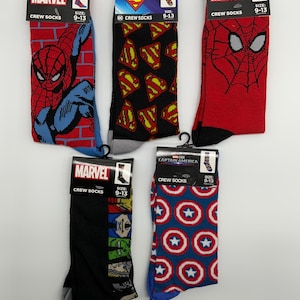 May include: Five pairs of crew socks featuring superhero designs. The socks include Spider-Man, Superman, and Captain America. Each pair is displayed on a black hanger with the Marvel or DC Comics logo and the size 9-13.