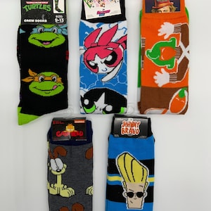 May include: Five pairs of crew socks featuring cartoon characters. Designs include Teenage Mutant Ninja Turtles, Powerpuff Girls, a character with a top hat, Garfield, and Johnny Bravo. Each pair is size 9-13.