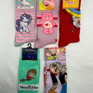 Puede incluir: Se muestran cinco pares de calcetines de personajes, cada uno con un diseño único. Los calcetines presentan personajes como un unicornio, un Care Bear, Rainbow Brite, Harry Potter y Popeye. Los calcetines son de varios colores, incluyendo rosa, rojo y verde.