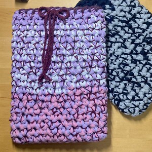 Peut inclure: Un sac rectangulaire fait à la main dans les tons de lavande, blanc et rose, avec une fermeture à cordon. À côté, un objet rond et plat, crocheté en bleu foncé et gris. Les deux articles sont en matériau épais et tissé.
