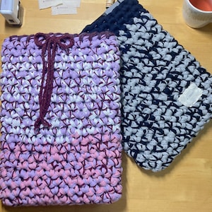 May include: Two crocheted bags, one in a purple, pink, and white color scheme with a burgundy drawstring, and the other in a black and gray color scheme.