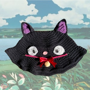 May include: A black crochet cat hat with white eyes, a pink nose, and a red bow. The hat has purple inner ears and a yellow bell. The hat is handmade and has a whimsical design.