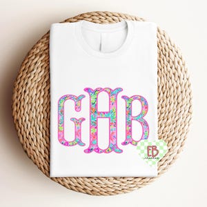 Puede incluir: Una camiseta blanca con un monograma floral en rosa, azul y verde, con las letras GHB. La camiseta está doblada sobre una estera redonda de mimbre.