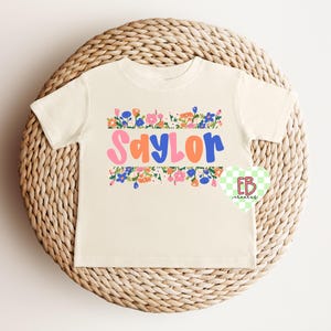 May include: A cream colored t-shirt with a floral border and the name "Saylor" in colorful letters.
