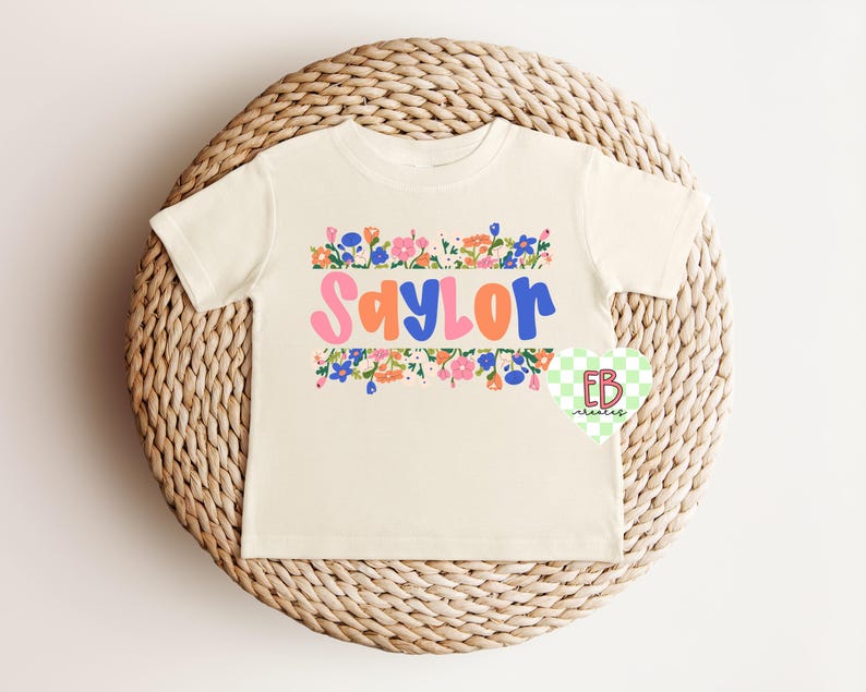 May include: A cream colored t-shirt with a floral border and the name "Saylor" in colorful letters.