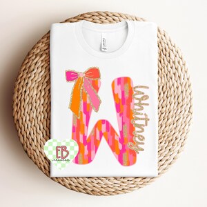 May include: A white t-shirt with a pink, orange, and gold glitter letter "W" with a pink and orange bow on top. The name "Whitney" is written in gold glitter next to the letter.
