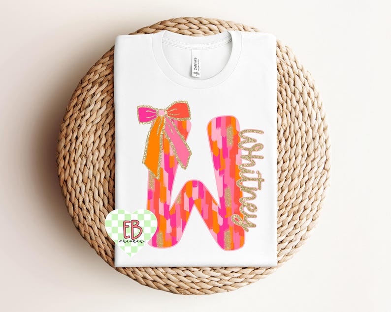 May include: A white t-shirt with a pink, orange, and gold glitter letter "W" with a pink and orange bow on top. The name "Whitney" is written in gold glitter next to the letter.