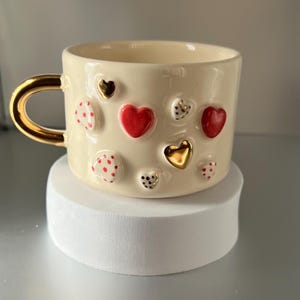 Op de afbeelding: Een witte keramische mok met een gouden handvat en rode en gouden hartvormige decoraties.