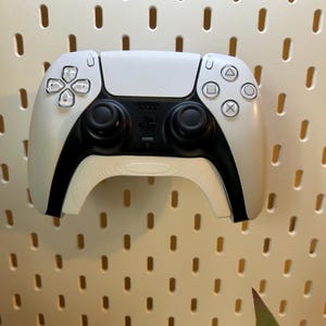 Könnte beinhalten: Ein weißer PlayStation 5 DualSense Wireless-Controller mit schwarzen Akzenten und Knöpfen. Der Controller hängt an einer Lochwand.