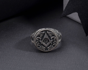 Könnte beinhalten: Ein silberner Ring mit einem verzierten Design, das einen Kompass und ein 'G' in der Mitte zeigt. Der Ring liegt auf einer dunkelgrauen Oberfläche.