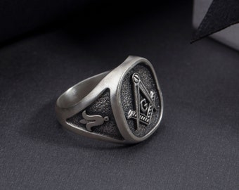 May include: A silver ring with a square-shaped design featuring a compass and square, a common symbol of Freemasonry.