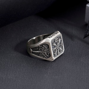 May include: A silver ring with a square face featuring a detailed design of a tree and a compass. The ring is on a black background.