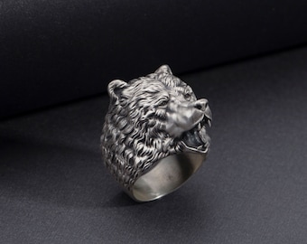May include: A silver ring with a detailed bear head design. The bear's mouth is open and its teeth are visible. The ring is on a black background.