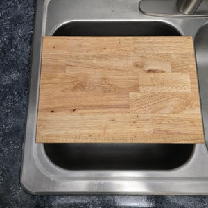 May include: A rectangular wooden cutting board with a light brown finish rests inside a stainless steel kitchen sink. The cutting board is made of multiple wood pieces joined together. The sink is set into a dark countertop.