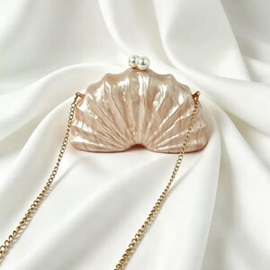 Pode incluir: Uma clutch em forma de concha perolada, com uma corrente dourada. A clutch é de cor pêssego claro e apresenta dois pequenos detalhes de pérolas brancas no topo. O fundo é um tecido branco e sedoso.
