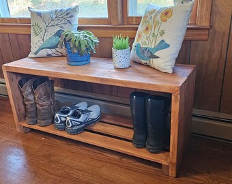May include: A stained wooden shoe bench with two shelves. The top shelf holds two decorative pillows with bird and floral designs, and potted plants. The bottom shelf displays various shoes and boots, including cowboy boots, sneakers, and rain boots.