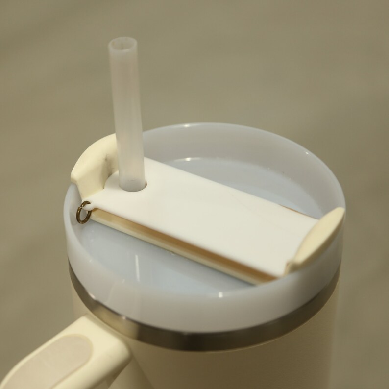 May include: A white travel mug with a lid that has a straw holder and a slide to cover the straw opening. The mug has a silver band around the middle.